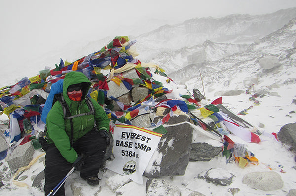 A photo of a plus-size woman sitting at Everest Base Camp in a blizzard. She is wearing a green jacket and black pants. Her face is almost completely covered by a face mask and glasses.